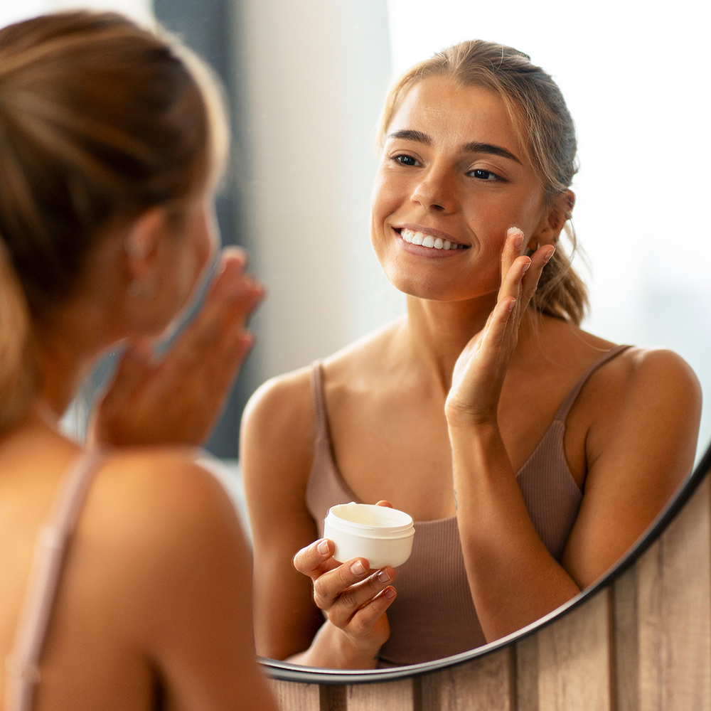 Femme bronzée s'appliquant une crème sur le visage devant un miroir